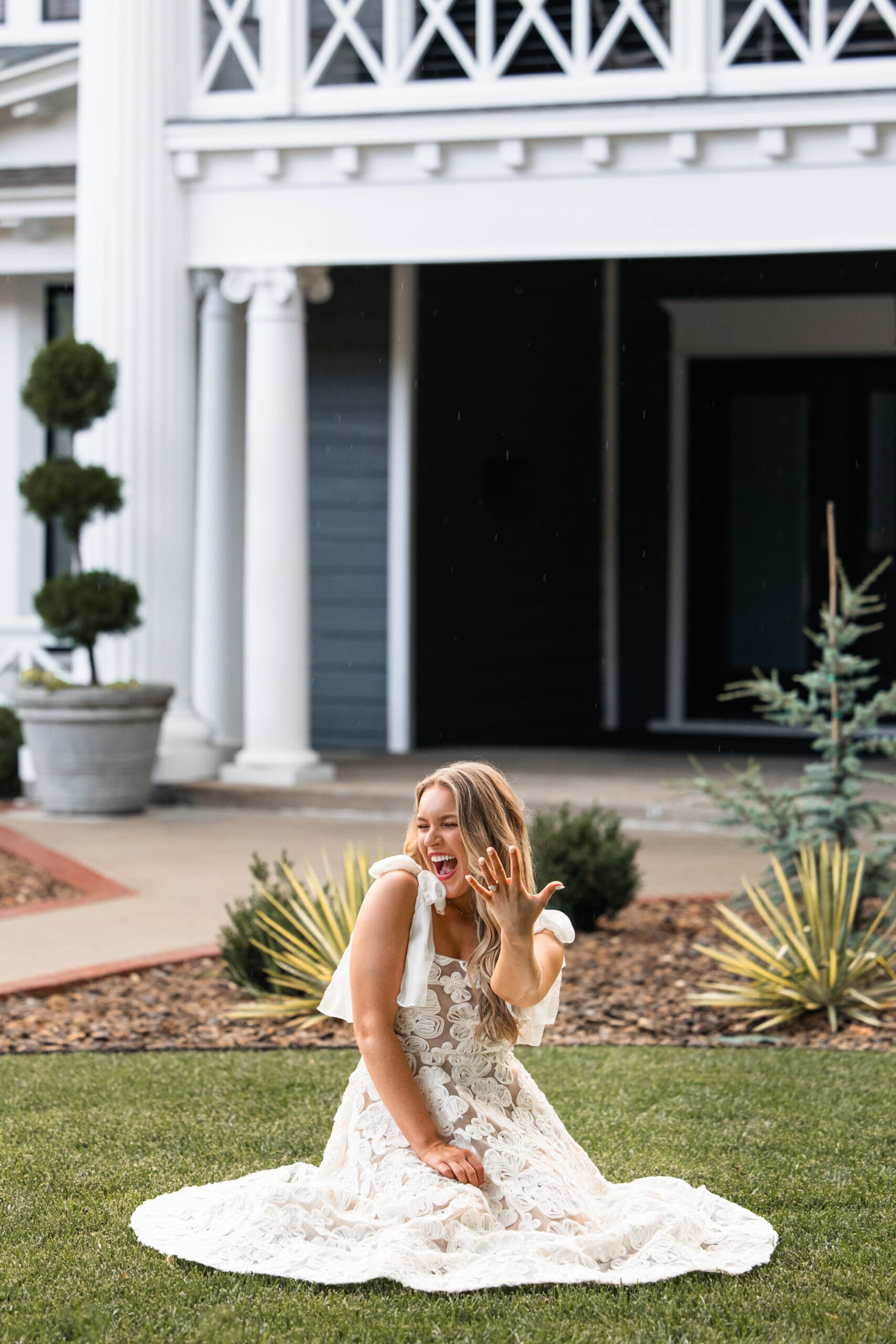 Bride sits in grass showing off her ring with a joyful expression. Wichita Engagement Photographer