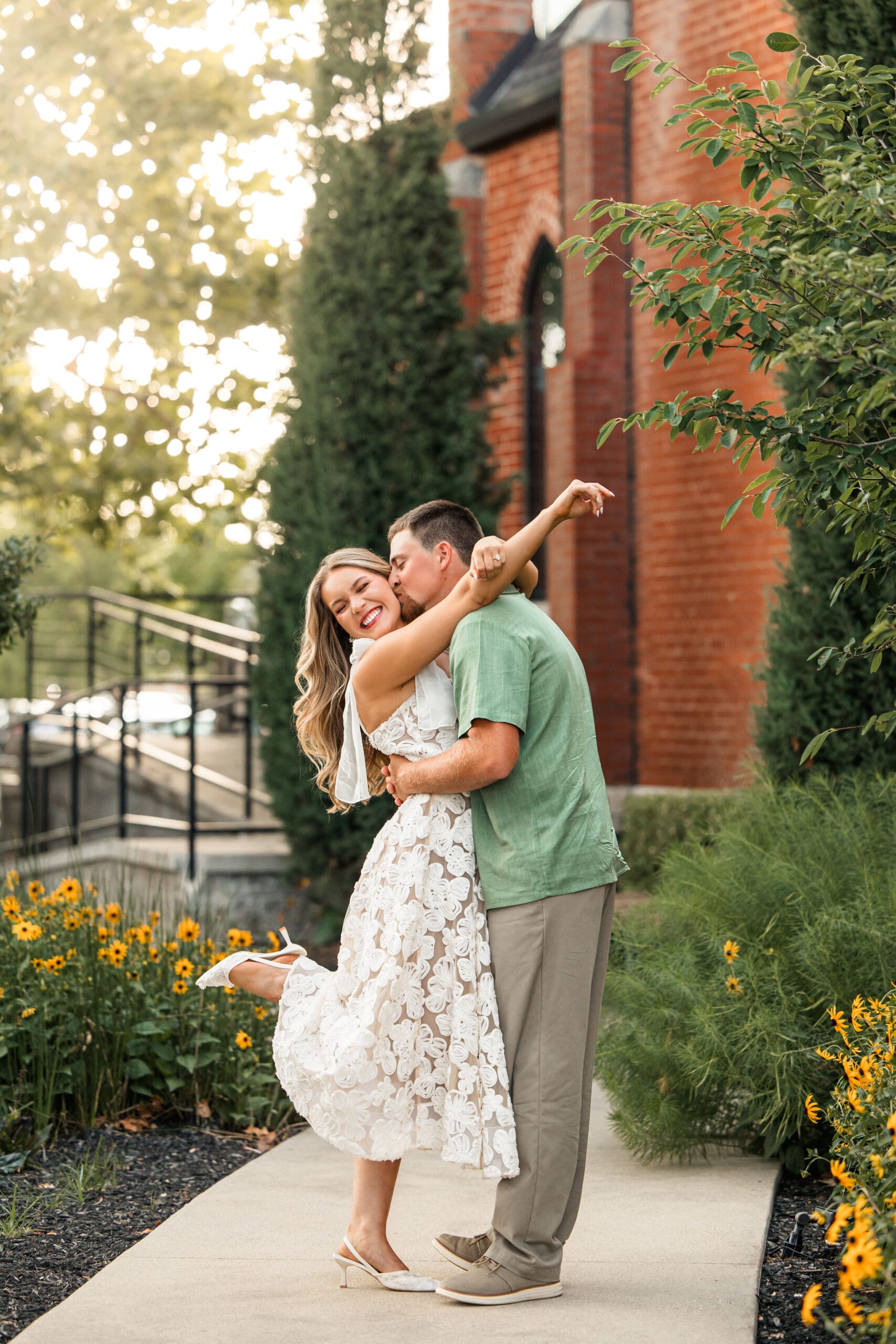 Full-body golden hour kiss surrounded by yellow flowers on a sidewalk, Wichita Engagement Photographer