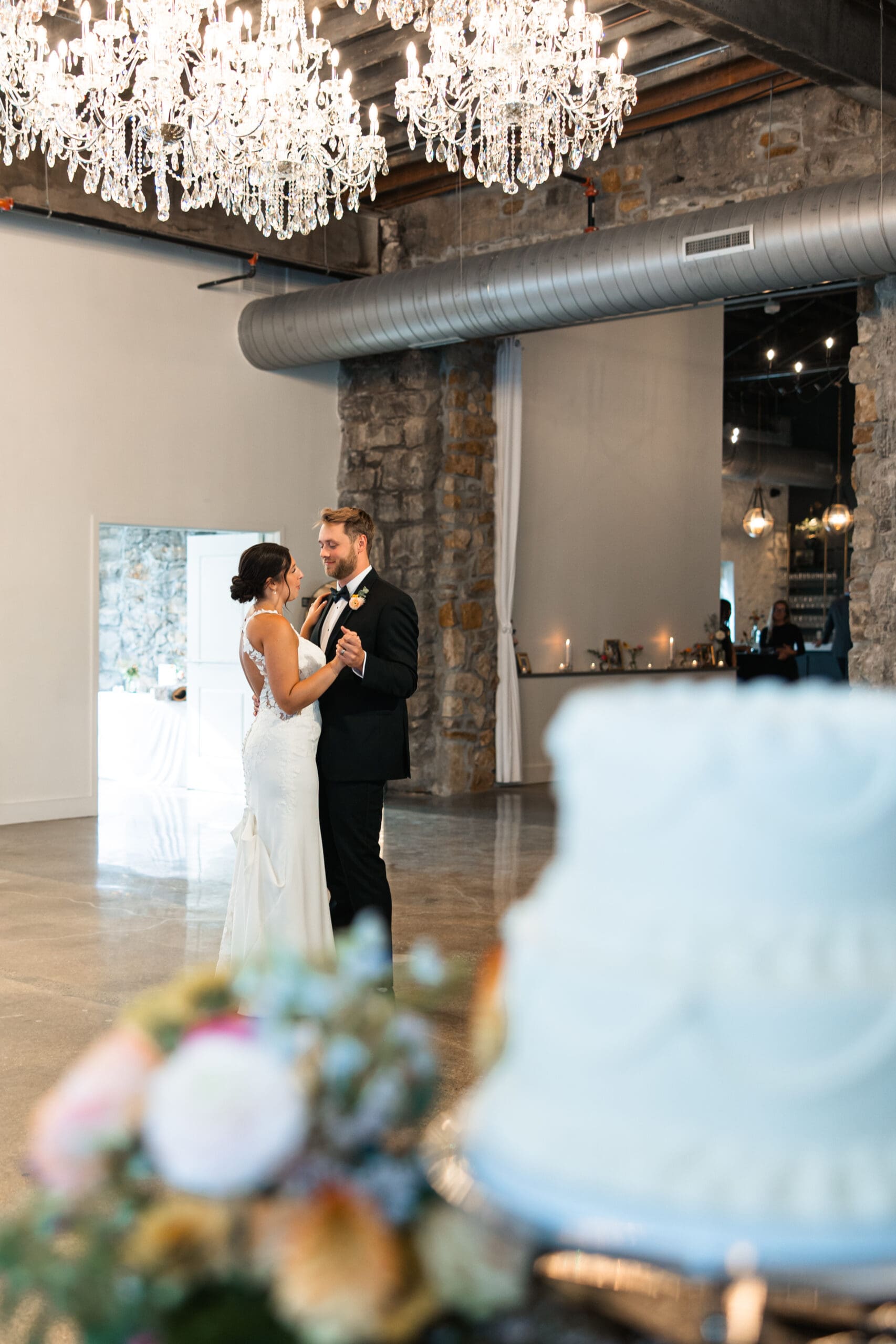 Bride and groom’s romantic first dance beneath chandeliers at Stone Manor on 79th, with the wedding cake softly in view.
