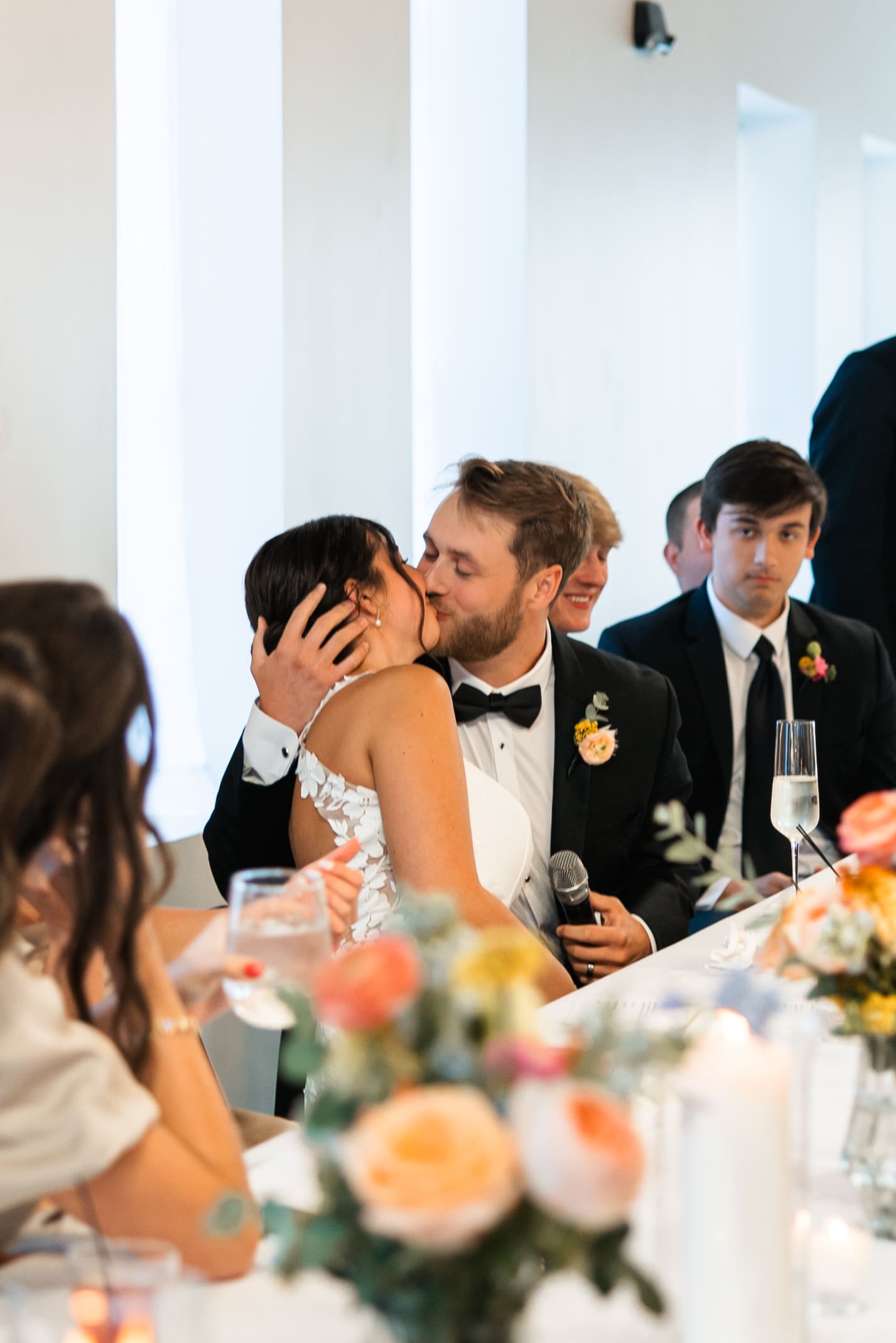 Bride and groom share a kiss during toasts at the head table, surrounded by colorful florals at Stone Manor on 79th in Overland Park.