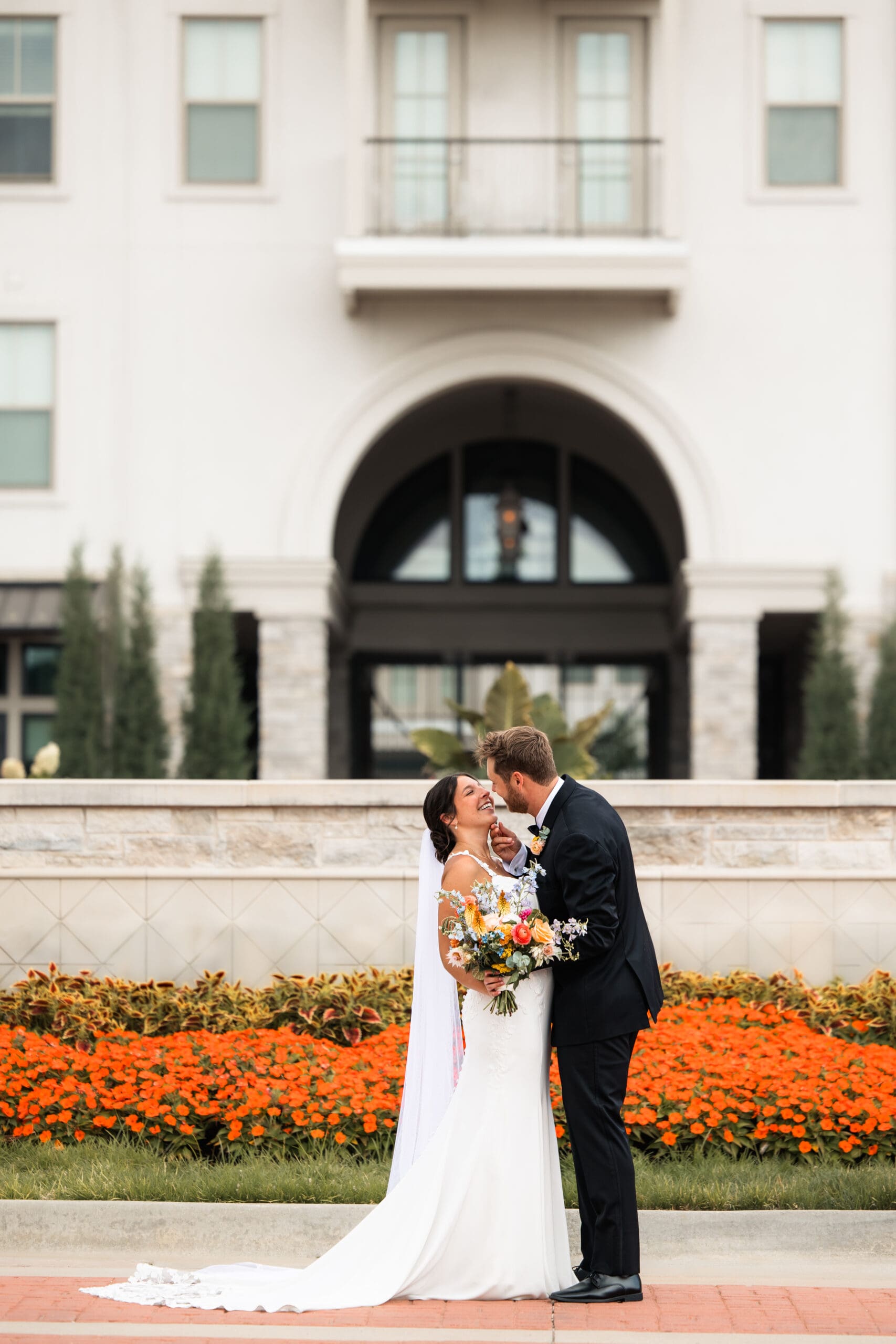Bride and groom kiss in front of The Kessler Residences, surrounded by blooming flowers, captured by high-end Kansas wedding photographer Briana Jewell Photography