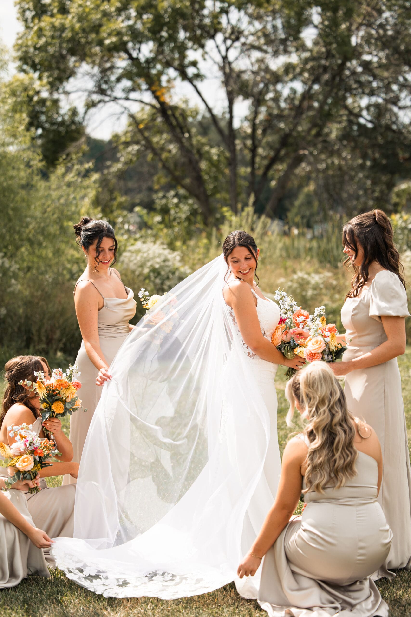 Bride surrounded by bridesmaids in champagne dresses, holding her veil in the sunshine at Meadowbrook Park in Overland Park, Kansas.