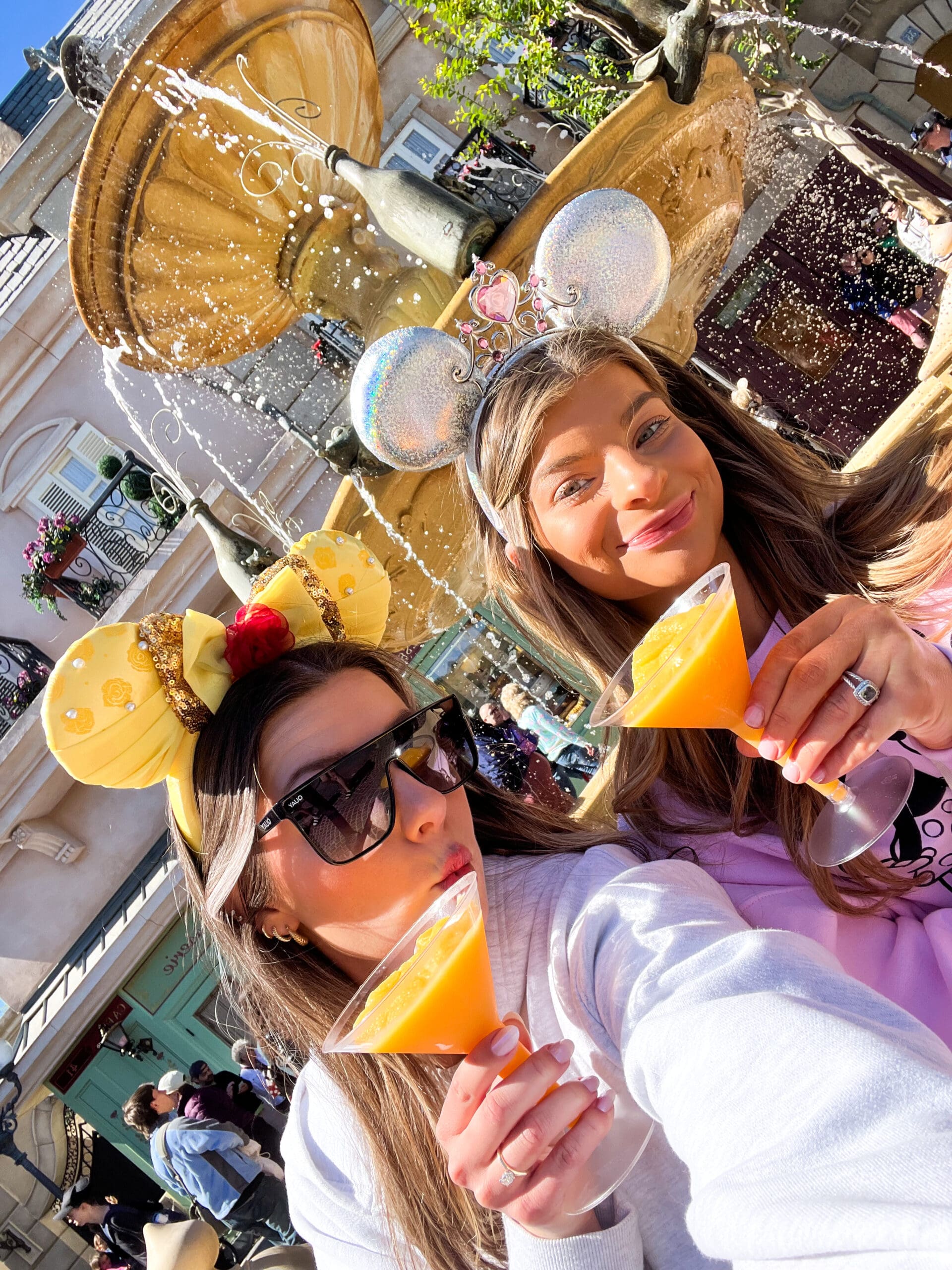 Two friends in sparkling Minnie Mouse ears sipping orange slush drinks at a fountain in Magic Kingdom Disney World – joyful, refreshing moment during Orlando Disney magic