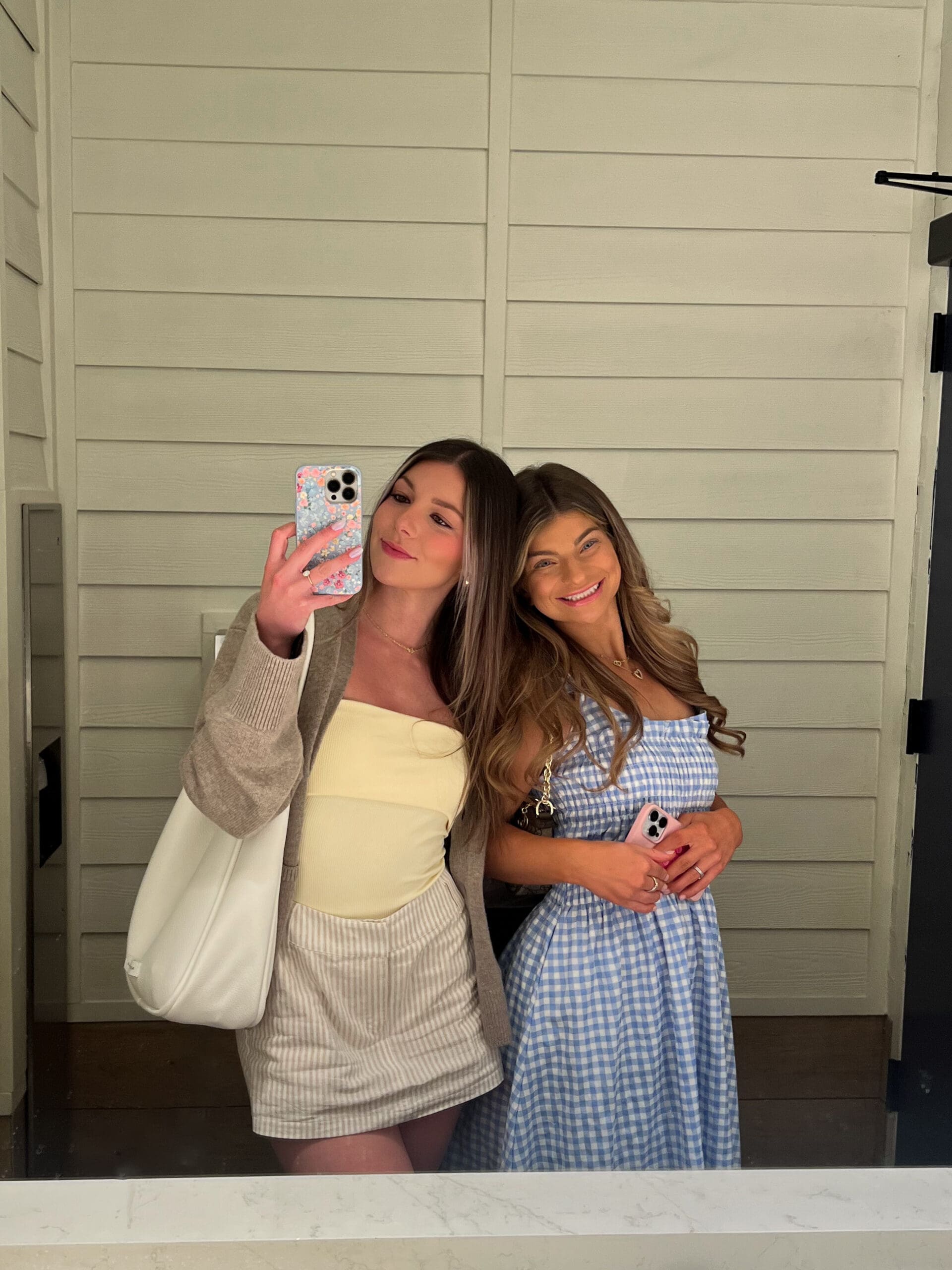 Two friends taking mirror selfie – one in white bodysuit and cardigan, the other in blue checkered dress – fun behind-the-scenes from Ave & Liv boutique Orlando shoot