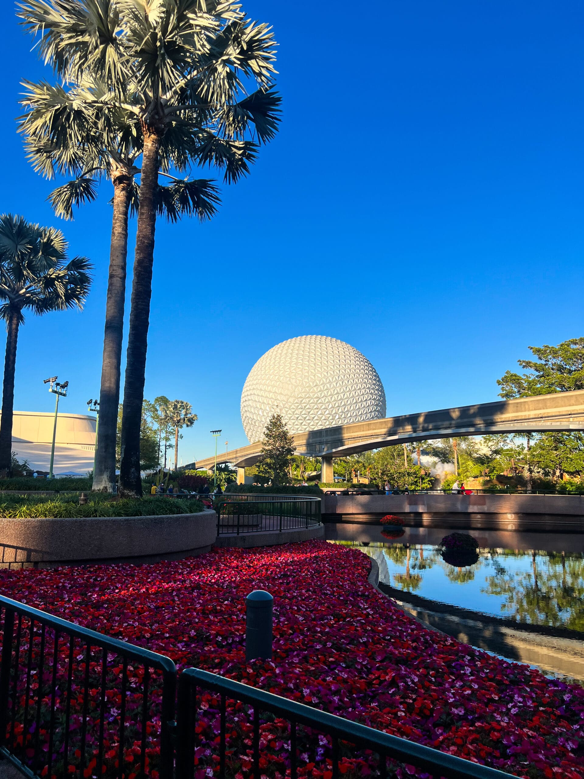 Iconic Spaceship Earth geodesic sphere framed by tall palm trees and vibrant red flower beds at Epcot Disney World – futuristic magic in Orlando sunset glow