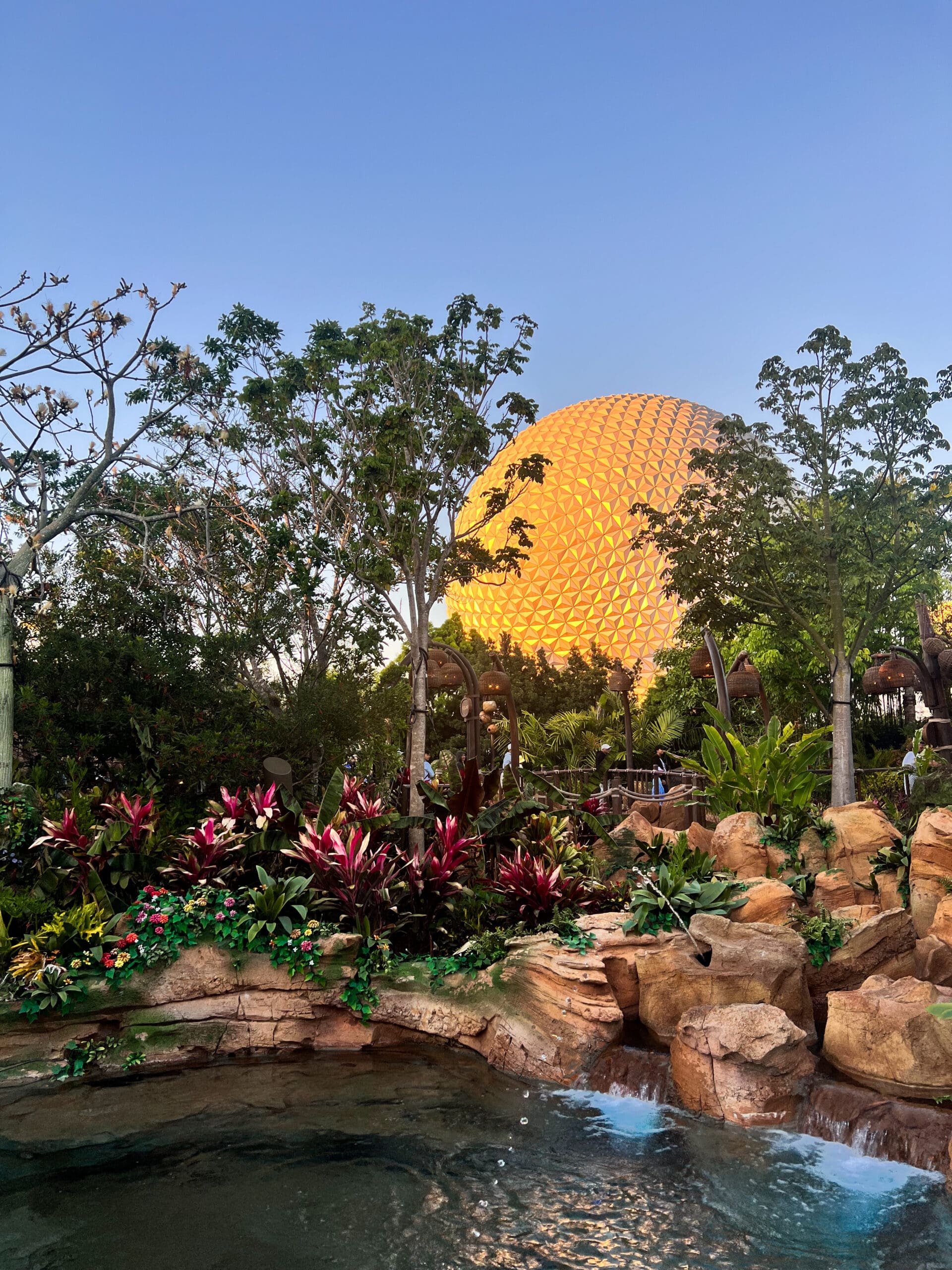 Spaceship Earth reflecting in water with palm trees and red flower pathway at Epcot Disney World – iconic futuristic landmark in Orlando sunset light