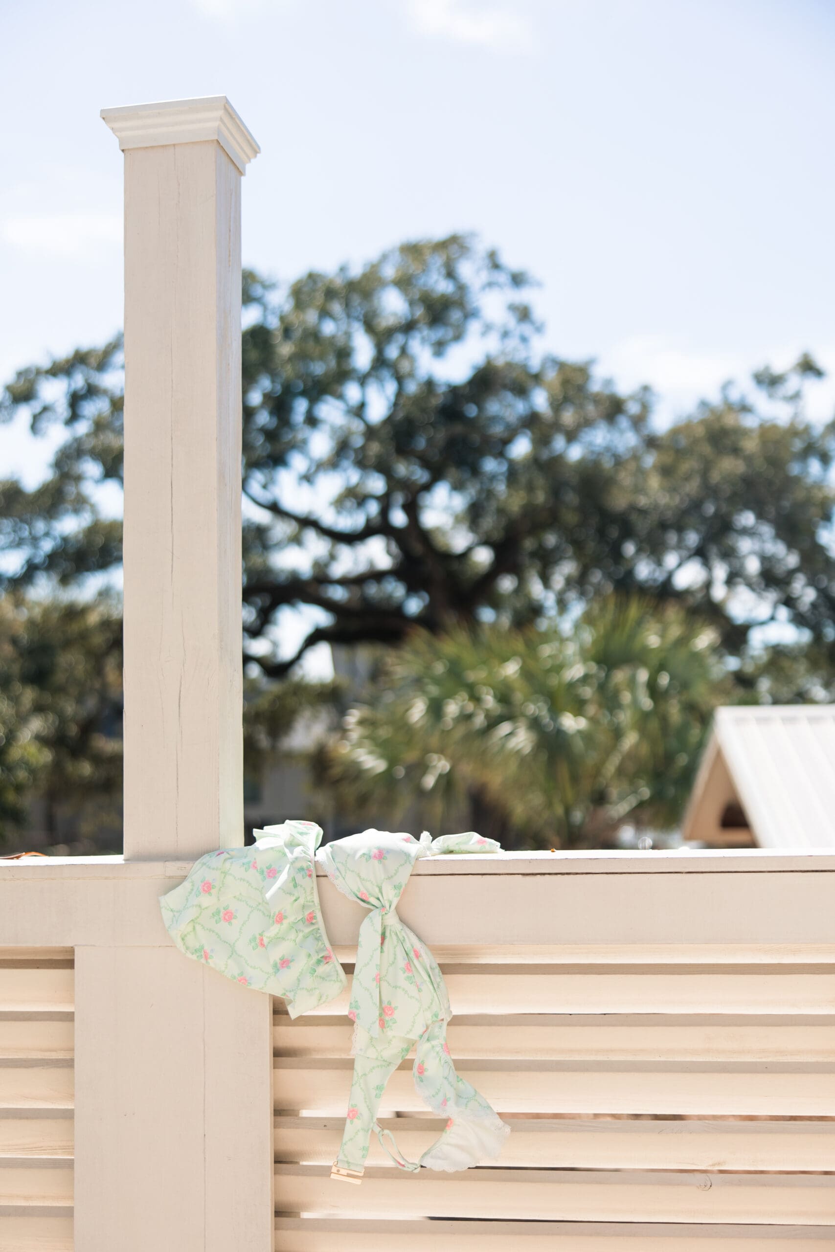 Light green floral bow from Ave & Liv’s new swimwear collection tied on a white railing during a Fairhope Alabama photoshoot, with oak trees and blue sky in the background