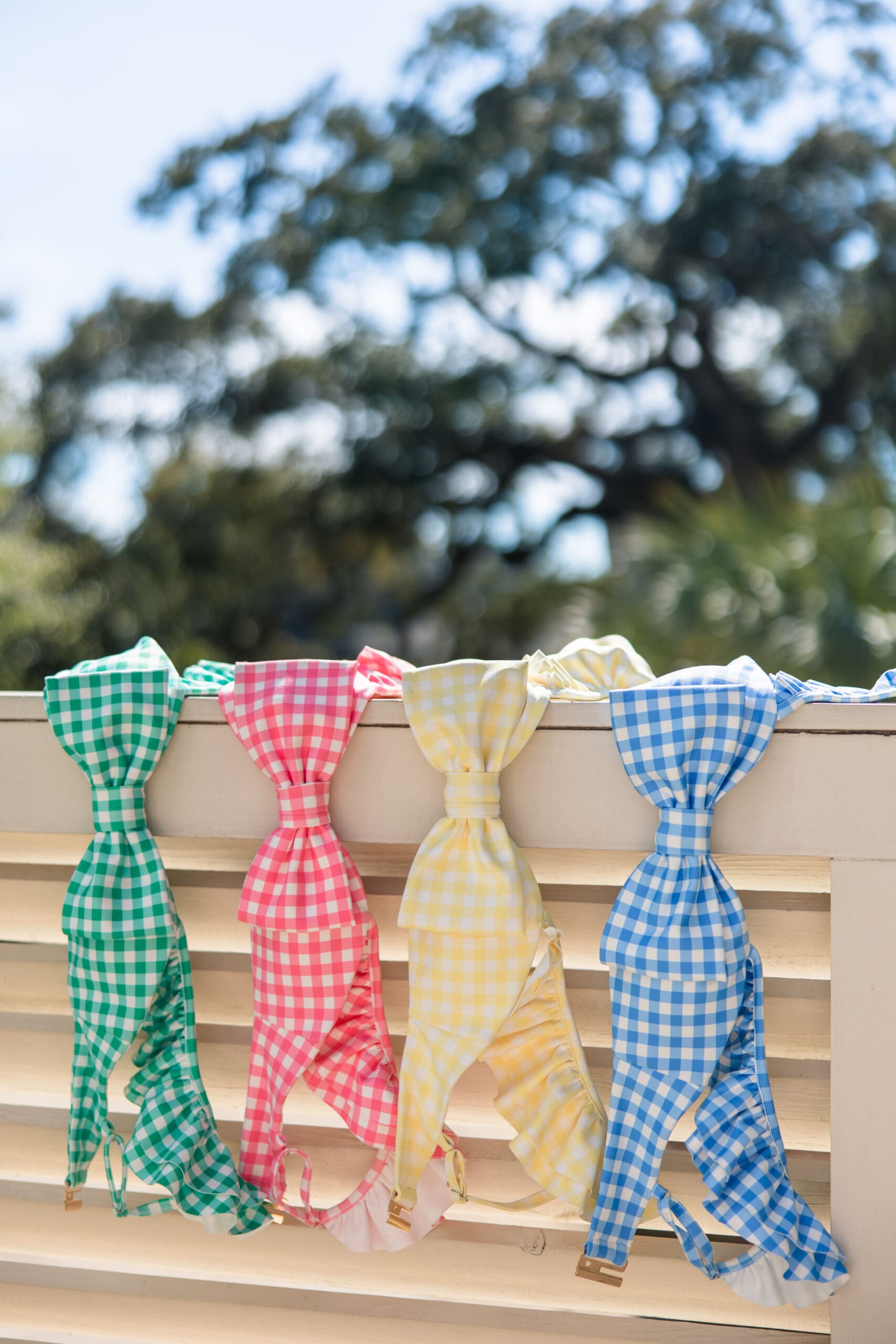 Four colorful gingham bikini tops in green, pink, yellow, and blue hanging on a white railing during the Fairhope Alabama photoshoot for Ave & Liv’s exclusive new swimwear launch