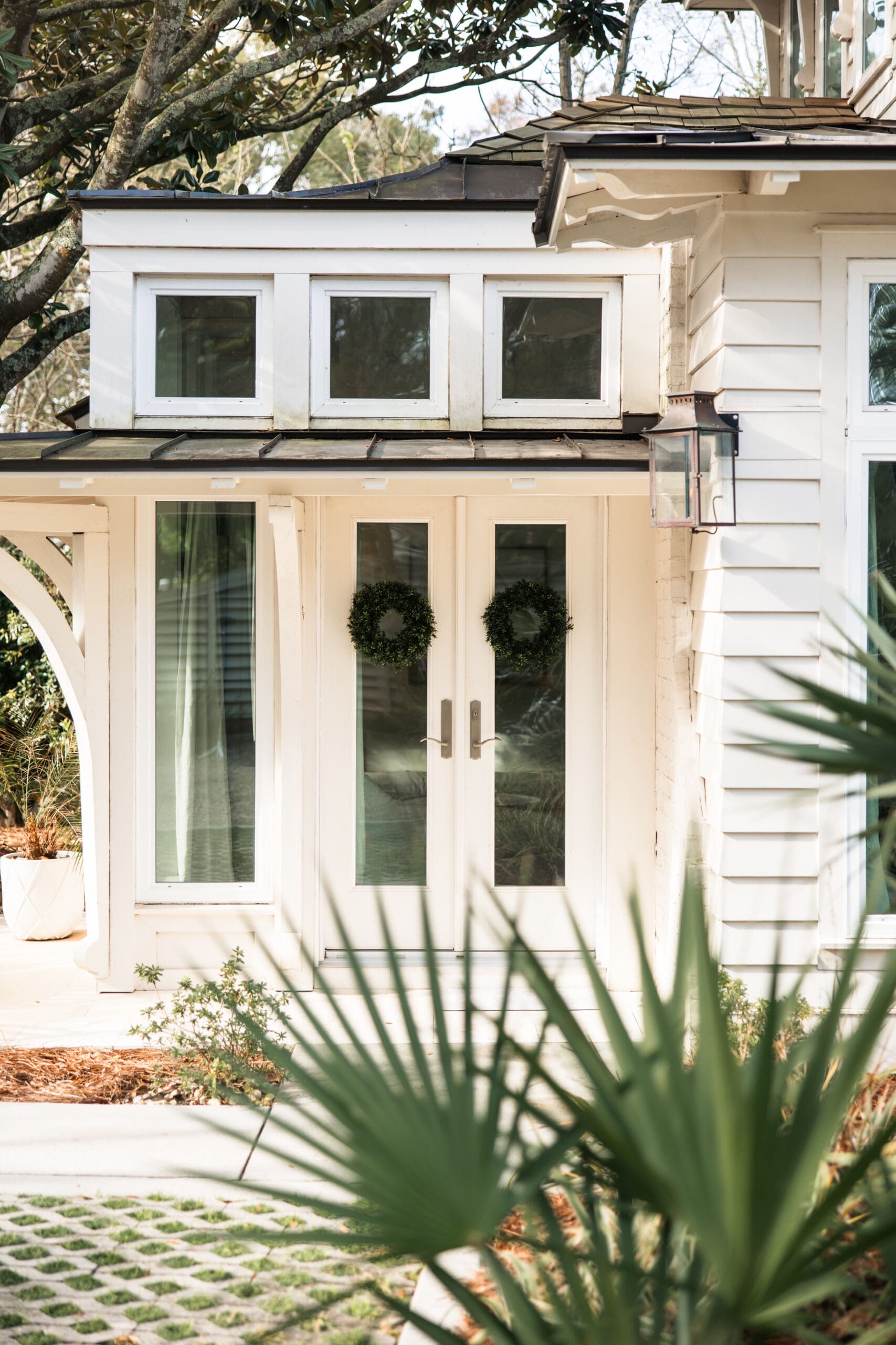 Charming white entrance of Lucia Bleu Fairhope Cottages with double doors and green wreaths during a Fairhope Alabama photoshoot, framed by palm leaves and oak trees