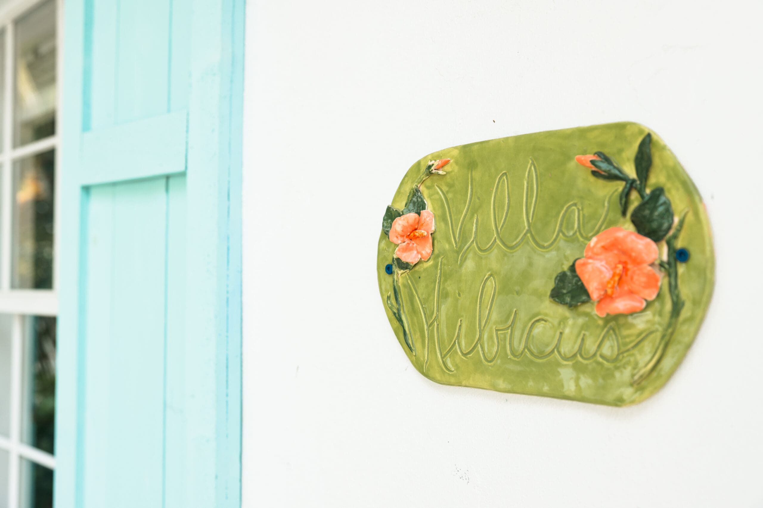 Hand-painted green ceramic "Villa Hibiscus" sign with orange hibiscus flowers on turquoise door at The Pink Palm Beach – cute detail for girly getaways