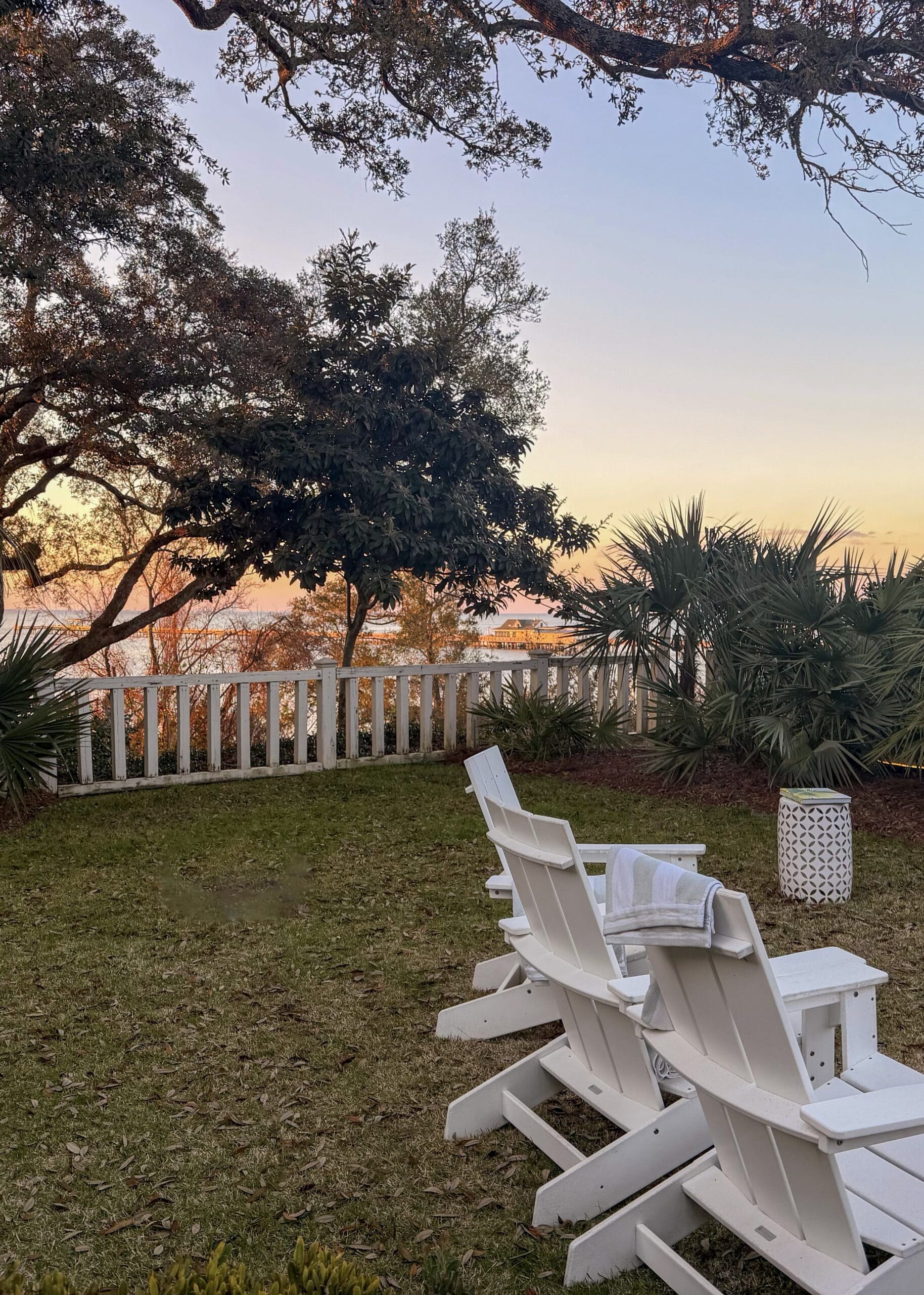 White Adirondack chairs on the lawn overlooking Mobile Bay at golden hour from Lucia Bleu Fairhope Cottages during a Fairhope Alabama photoshoot, with oak trees and palm plants framing the dreamy sunset view
