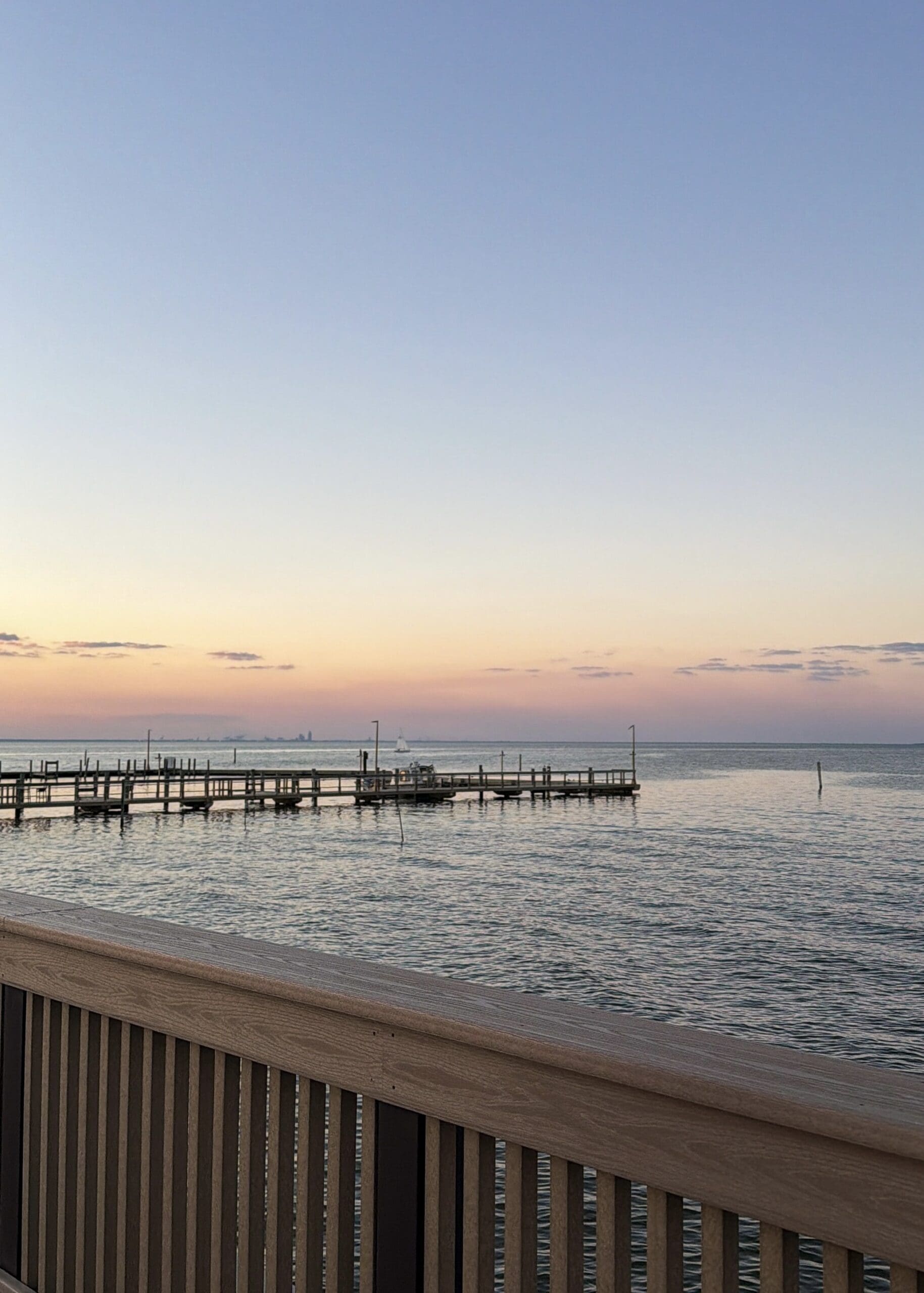Peaceful view of the wooden pier and calm water at sunset in Fairhope, Alabama during a Fairhope Alabama photoshoot, with a sailboat in the distance and soft pink and blue sky
