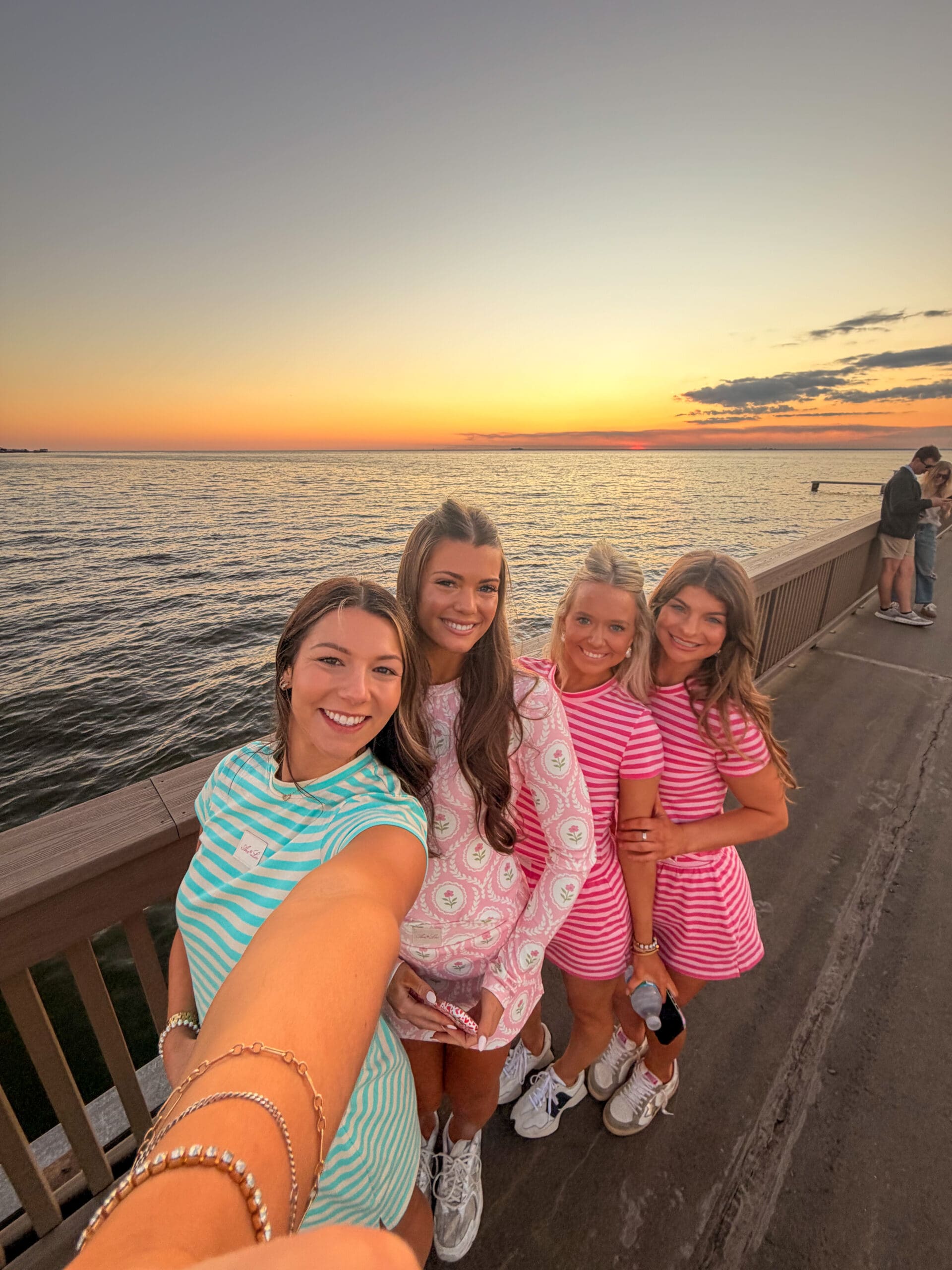 Four smiling women taking a sunset selfie on the Fairhope Municipal Pier during a Fairhope Alabama photoshoot, with Mobile Bay and a vibrant orange sky in the background